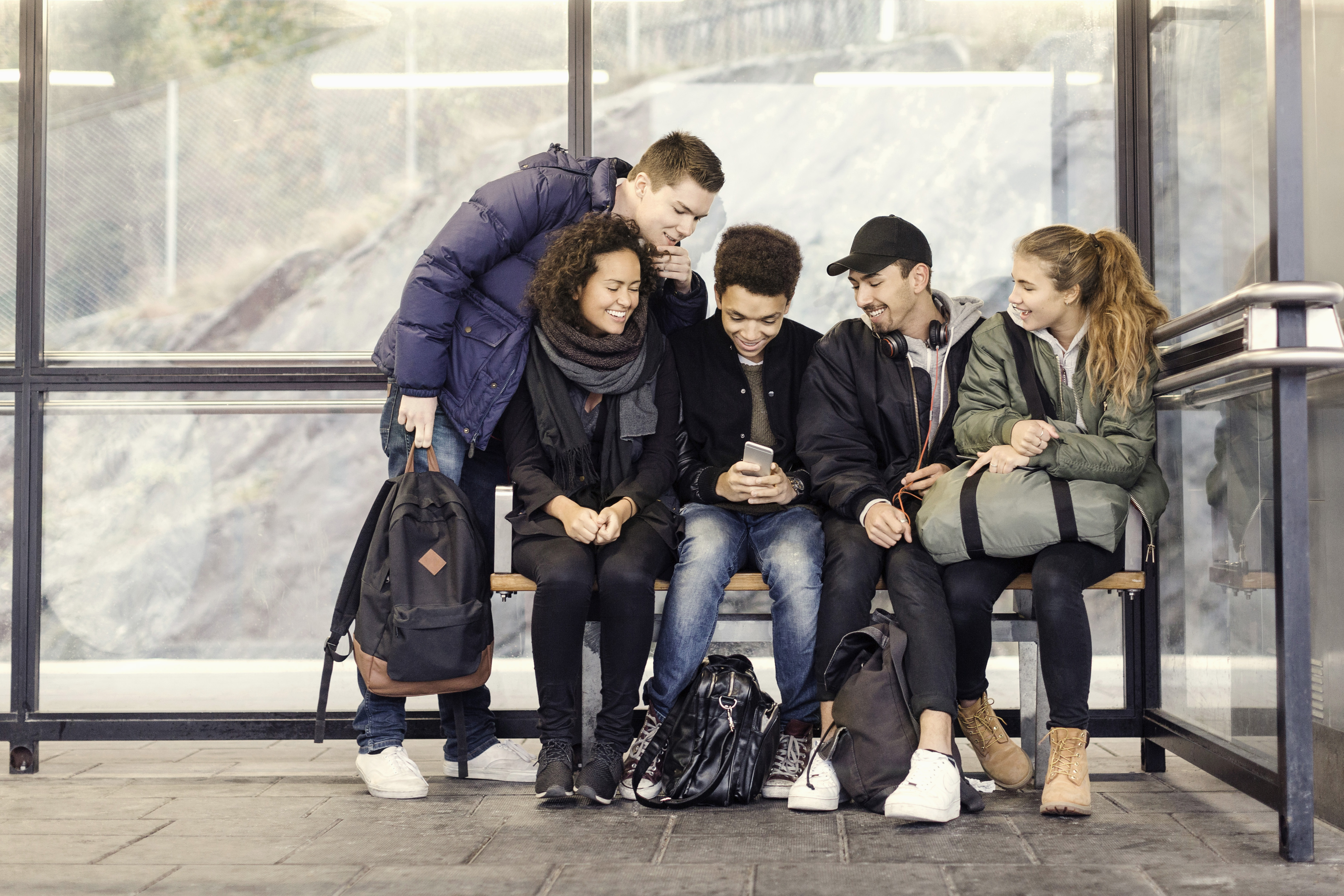 En grupp ungdomar sitter på en busshållplats, ler och tittar på en mobiltelefon